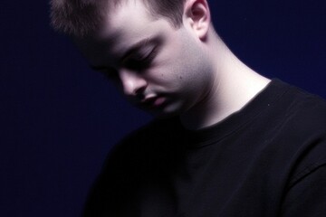Thoughtful Young Man in Dark Shirt Against Dim Blue Background Contemplating with Closed Eyes