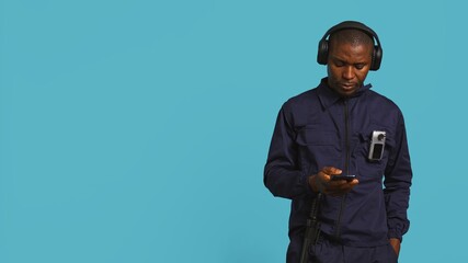 Security guard equipped with bodycam listening music, taking break from job shift. Portrait of bodyguard walking around perimeter with headphones on while at work, studio background, camera B