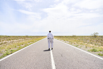 Obraz premium Man seen from behind walking towards the horizon in the middle of a quiet empty road. The scene conveys calm and tranquility.