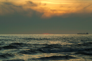 Obraz premium A serene seascape capturing the golden rays of a setting sun piercing through clouds above gentle ocean waves, with a distant ship on the horizon. 
