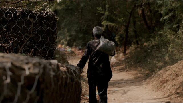 A handheld long shot of an old Indian crazy man with a white plastic bag on his shoulders walking away on a rural path surrounded by trees and vegetation, with his back towards the camera