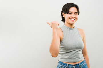 Young latin woman smiling and pointing to the side with her thumb while standing in a studio with a gray background