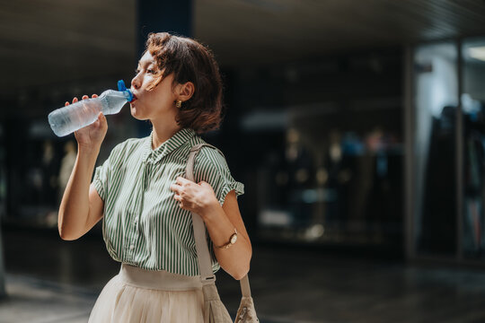 Young woman in green striped shirt drinking from a water bottle. Captured in an urban environment, highlighting hydration and refreshment.