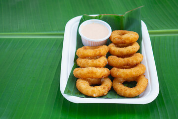 Golden onion rings served with sauce in biodegradable tray on green leaf.