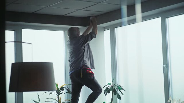 Professional male installer in uniform stands on ladder and screws surveillance camera to the ceiling in office room using tools. Monitoring and tracking. CCTV system, security and privacy concept.