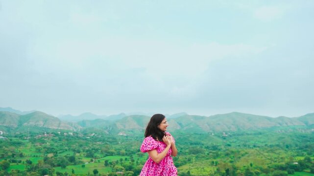 Portrait of beautiful teenager girl wearing pink dress looking at green hills in distant at Rayta, Udaipur, India. Stylish fashion influencer standing outdoors. 