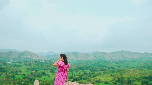 Portrait of beautiful teenager girl wearing pink dress looking at green hills in distant at Rayta, Udaipur, India. Stylish fashion influencer standing outdoors. 