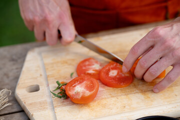 Close up of man holding sharp knife, slicing juicy red tomato. Preparing vegetables for an outdoor barbecue.