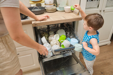 A mother woman and child unload a dishwasher in a home kitchen. Baby helps parents with household...