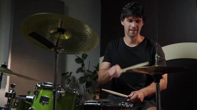 A young man sits behind a drum set in a home studio and plays with two drumsticks. The drums are green and black, and there are two cymbals. The drummer is wearing a black t-shirt, and he is looking