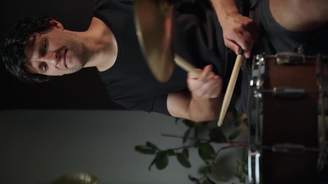 A young man plays drums with passion, his face illuminated by the light of the drum set. He is dressed casually in a black shirt and shorts. The room appears to be a home studio.