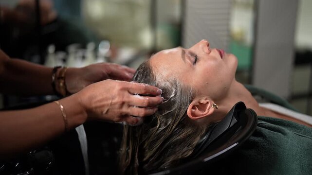 Close-up hands of unrecognizable hairstylist foaming shampoo on long blonde hair of beautiful woman. Professional hairdresser washing head of female client in beauty hairdressing salon, slow motion.