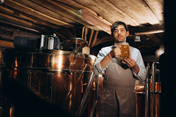 Happy brewer. Happy young male brewer in apron holding glass with beer and looking at it with smile while standing in front of metal containers