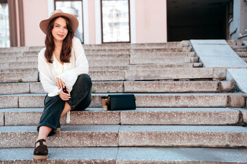 Young beautiful smiling hipster woman in trendy summer white blouse and jeans. Carefree woman posing in the street in sunny day. Positive model outdoors. Cheerful and happy. Sits at stairs. In hat