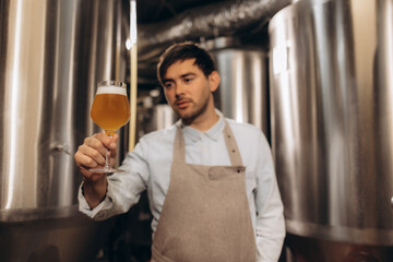 Bearded professional brewer holding clipboard, examining freshly made beer in a glass. Beer craftsman working at his microbrewery holding up beer mug. Manufacturing, food and drink industry concept