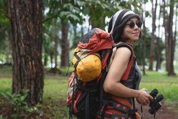 Portrait of Asian female backpacker holds binoculars to view natural scenery and birds while hiking...