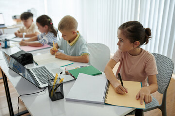 Fototapeta premium Group of children sitting at table in classroom writing in their notebooks with pencils. Laptop and stationery arranged on table while children concentrate