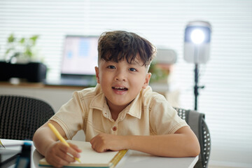 Caucasian child smiling while writing in notebook at home office with blurry background of computer and light source offering bright, cozy atmosphere