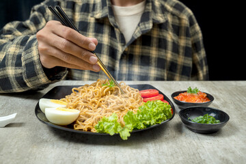 A man is eating a plate of instant noodles with a fork and chopsticks