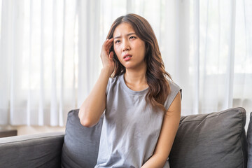 Young asian woman experiencing a headache sitting on a sofa with a pained expression, serious mood, health, wellness, stress, headaches and everyday struggles, pain and importance of self-care at home