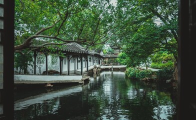 Fototapeta premium A view through a window of a tranquil Chinese garden. Lush greenery frames a serene pond with a white pavilion in the background.