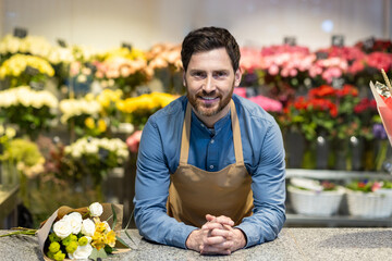 Smiling male florist in apron leaning on counter with bouquet surrounded by colorful flowers in flower shop. Person showcasing beautiful floral arrangements and customer service in bloom-filled store