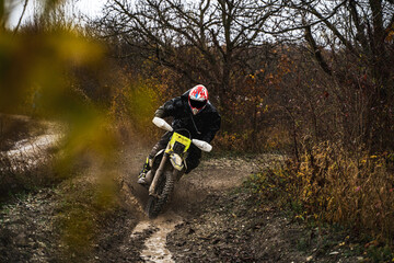 Obraz premium Professional motorcyclist in full moto equipment riding crops enduro bike, enduro driver rides through puddles in cloudy weather, motocross racer in a wet and muddy terrain, motorcycle race