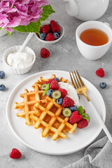 Delicious Belgian Waffle with whipped cream, fresh berries and mint on a white plate on a gray concrete background. Summer breakfast.