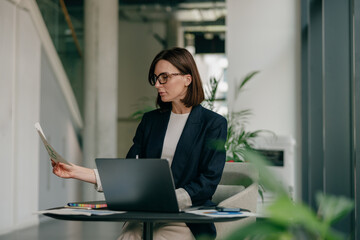 Focused businesswoman reviewing documents at office workspace with laptop and plants