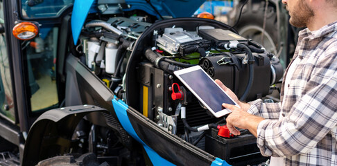 Mechanic diagnoses tractor engine operation using digital tablet. Blank screen of digital tablet in hands in front of machinery engine.