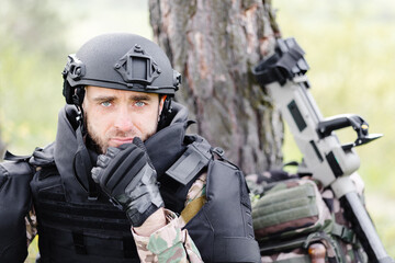 A man in a military uniform and a bulletproof vest sits in the forest near a metal detector and a...
