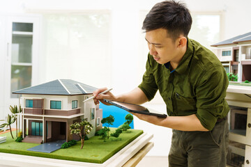 Asian businessman works alone at his desk, surrounded by a laptop, paperwork, tablet, and calculator, focusing on home and land mortgage agreements in a modern house model.