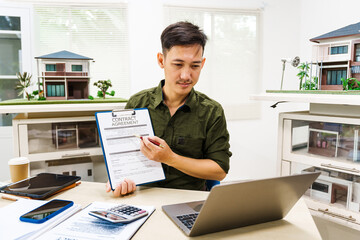 Asian businessman works alone at his desk, surrounded by a laptop, paperwork, tablet, and calculator, focusing on home and land mortgage agreements in a modern house model.