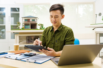 Asian businessman works alone at his desk, surrounded by a laptop, paperwork, tablet, and calculator, focusing on home and land mortgage agreements in a modern house model.