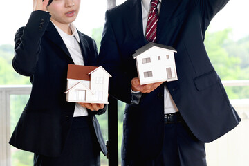 住宅模型を持ちながら困り顔のスーツ姿の男女のビジネスパーソン