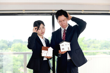 住宅模型を持ちながら困り顔のスーツ姿の男女のビジネスパーソン