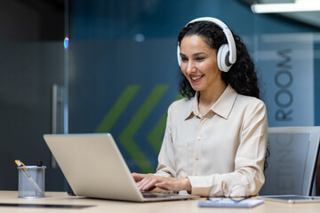 Smiling businesswoman wearing headphones working on laptop during online meeting in modern office...