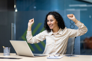 Happy businesswoman raising arms in success while working on laptop in modern office. Professional...