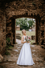 Cesis, Latvia - July 12, 2024 - A smiling bride in a white gown stands holding a bouquet in a stone archway, with a rustic and historic background.