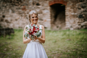 Cesis, Latvia - July 12, 2024 - A joyful bride in a white dress holds a bouquet of red and pink flowers, standing outdoors near an ancient stone wall.