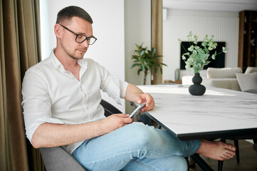 Portrait of man engaging with smartphone sitting at white marble countertop on kitchen, wearing...