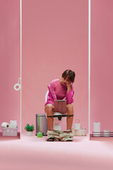 Obraz premium Young girl in pink clothes, with short hair sitting on toilet and looking on tablet with bored face against pink background. Concept of sport, game, match event, human emotions