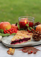 Piece of blueberry pie on a plate with autumn decorations, candle and red flowers. Light grey background with copy space. Cozy autumn food. 