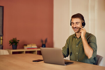 A smiling man having a conversation with a client over headphones with a microphone, during an online meeting.