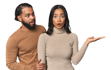 Young Latino couple in studio impressed holding copy space on palm.