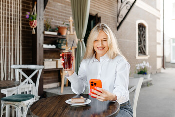 young beautiful blond caucasian woman sitting at table in outdoor terrace of cafe in summer day with chocolate cheesecake and strawberry cocktail, businesswoman looking at smartphone and smiling