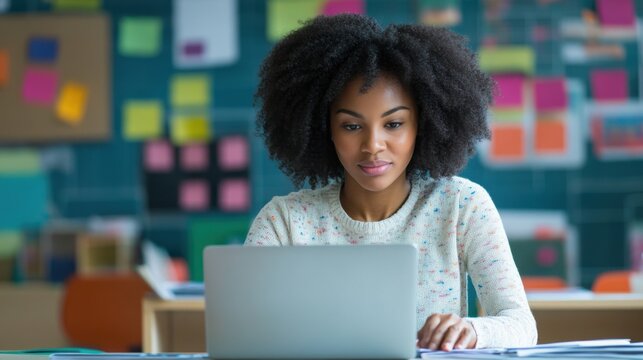 A young African American teacher sitting at her desk in a modern classroom, working on a laptop, preparing lesson plans, and utilizing digital resources to enhance her teaching methods