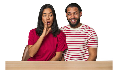 Seated young Latino couple at table is saying a secret hot braking news and looking aside