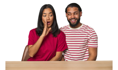 Seated young Latino couple at table is saying a secret hot braking news and looking aside