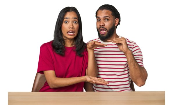Seated young Latino couple at table shocked and amazed holding a copy space between hands.